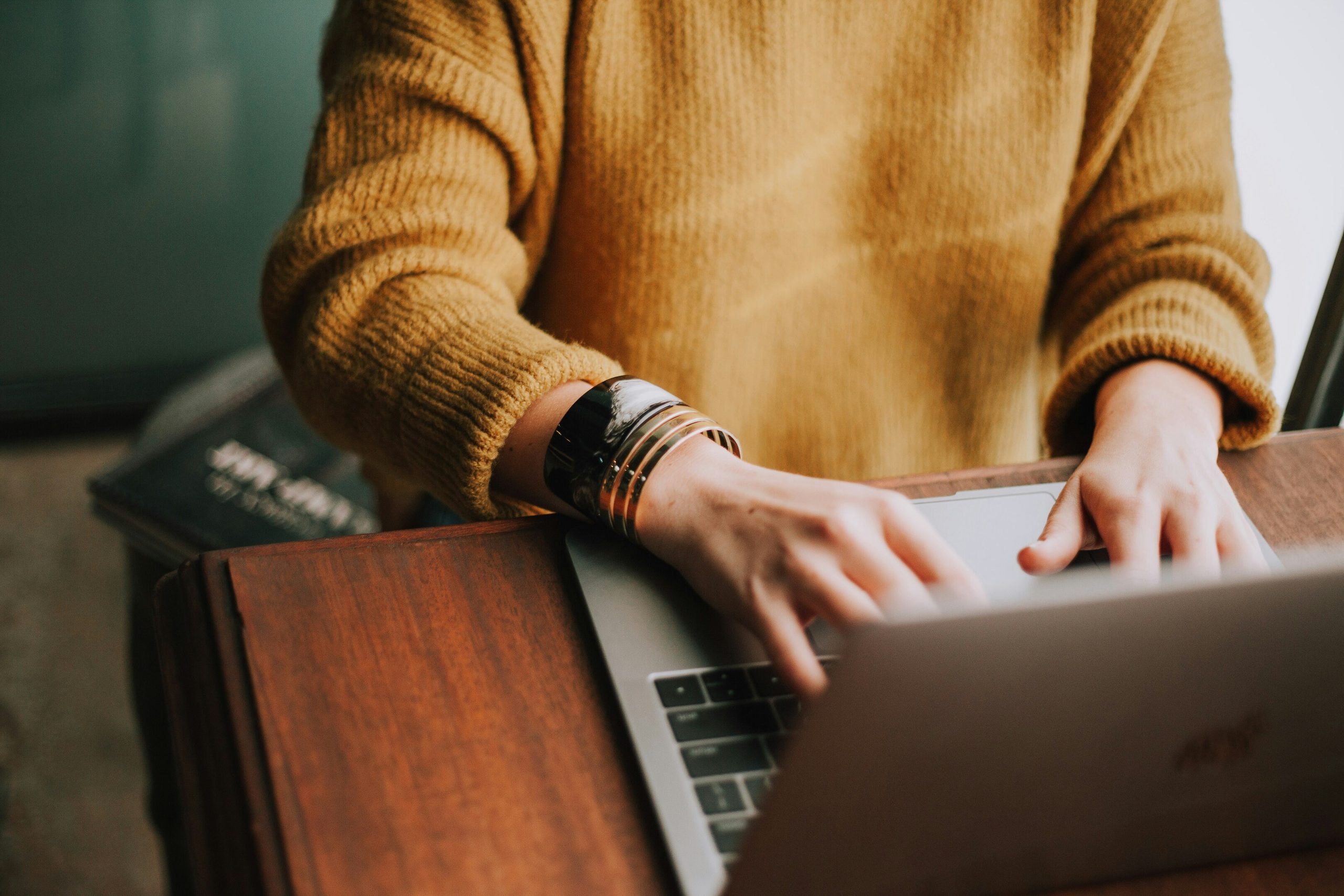 Close-up of hands wearing a bracelet, typing on a silver laptop at a wooden table.