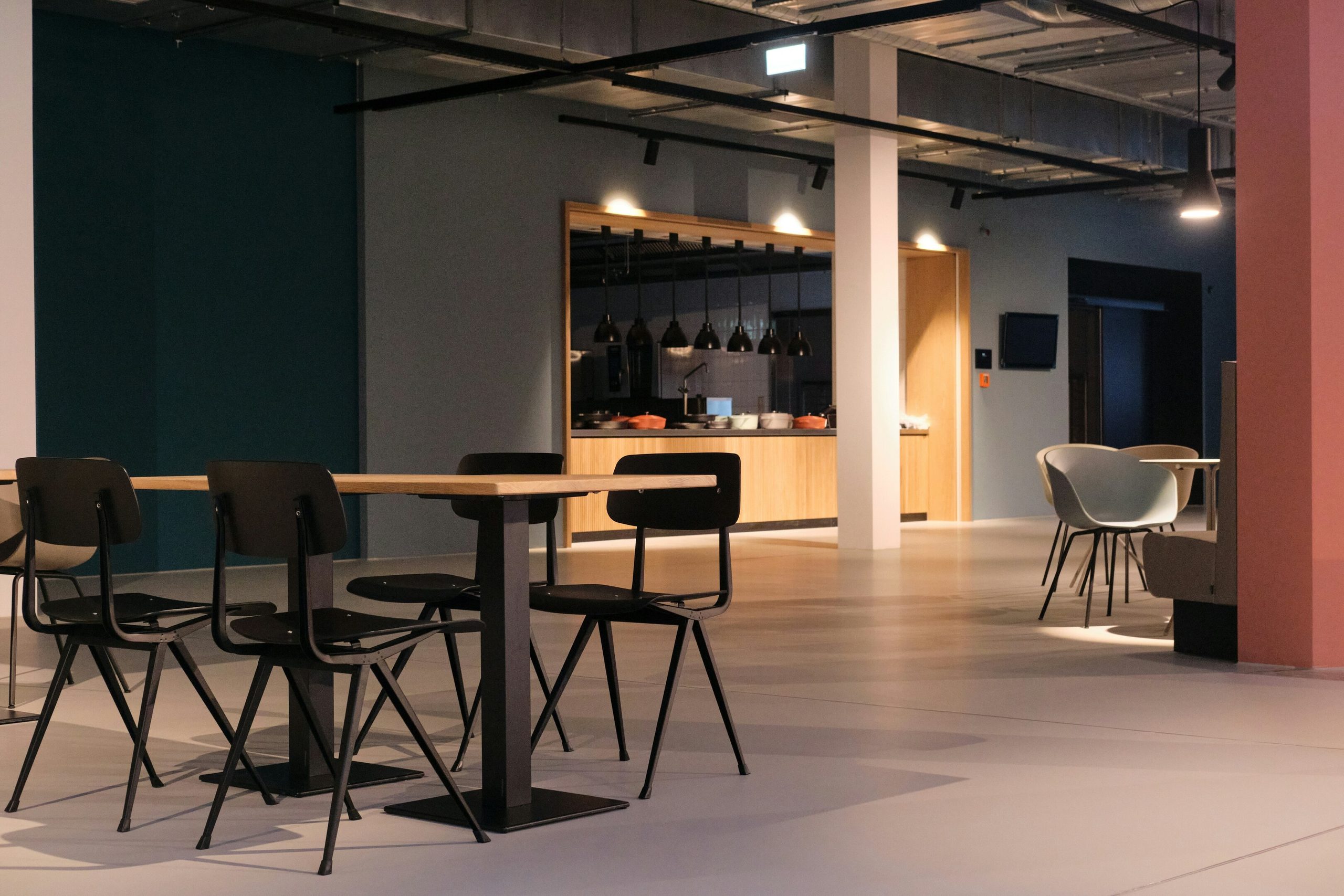 Modern communal dining area with multiple tables and chairs, and a kitchen/bar in the background.