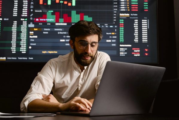 Man in glasses uses a laptop in front of a large screen displaying stock market charts.