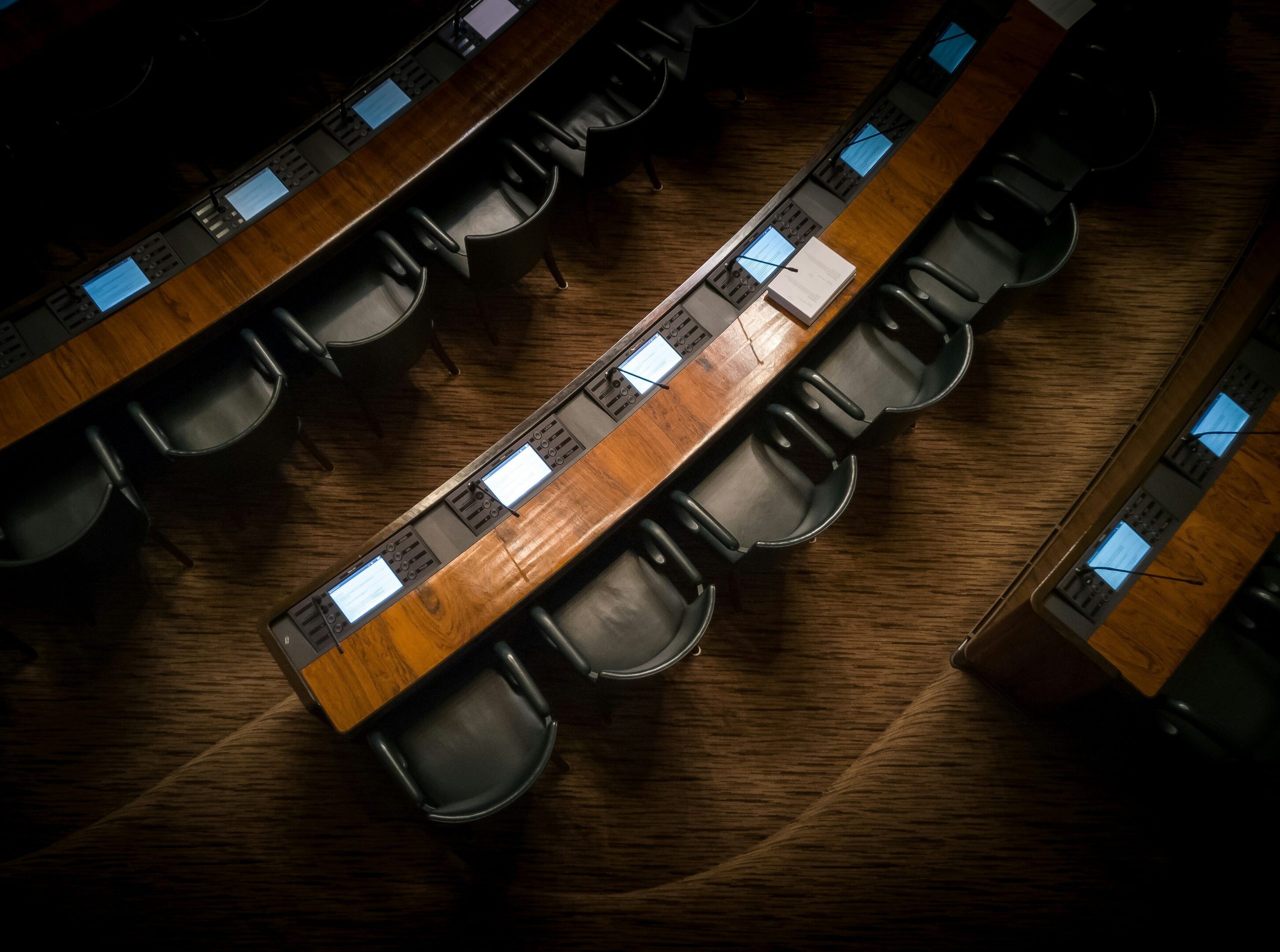 Overhead view of empty, curved conference desks with chairs and glowing screens in a dark hall.