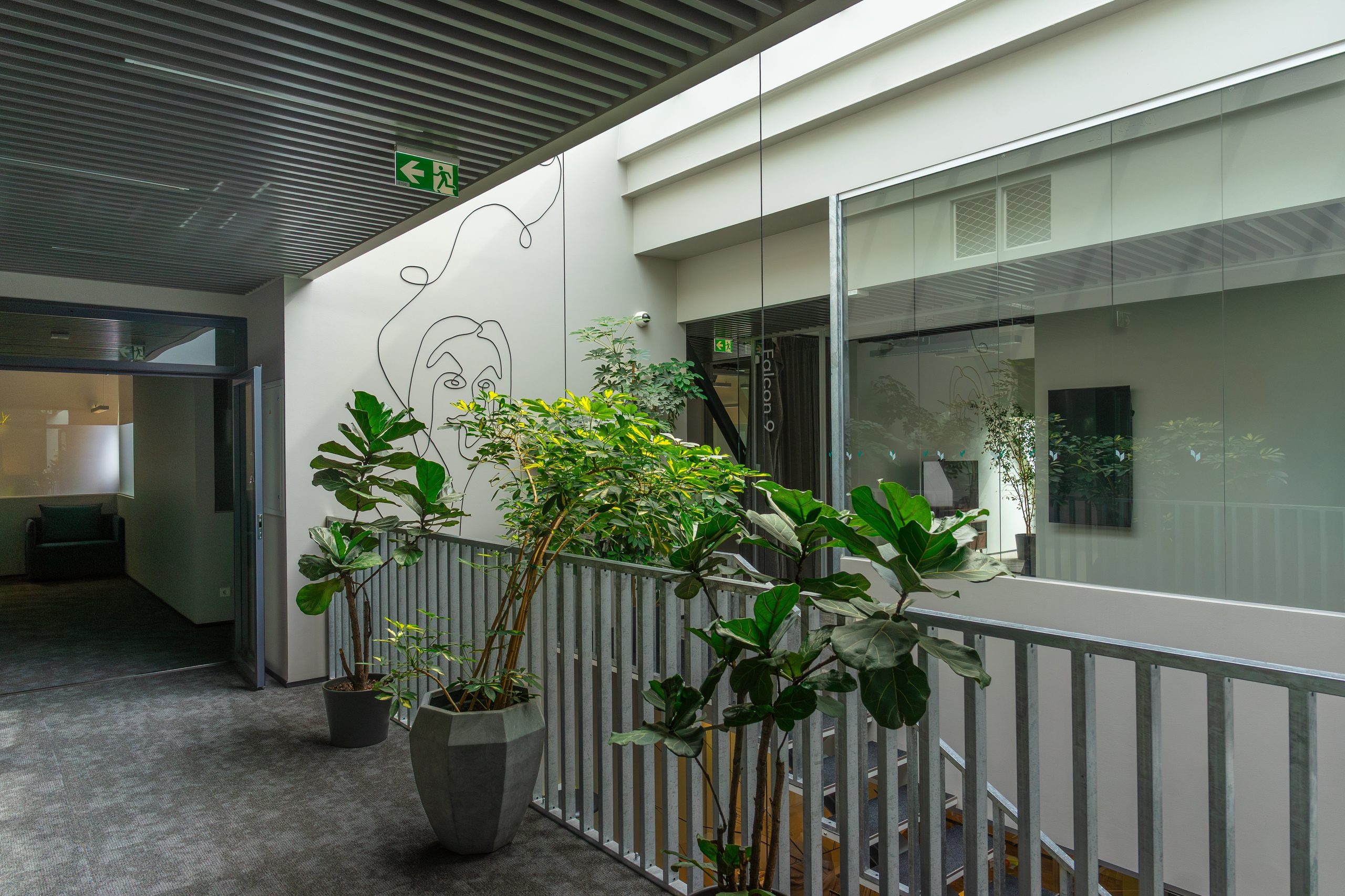 Modern office hallway with grey carpet, a metal railing, green potted plants, and abstract line art on a white wall.