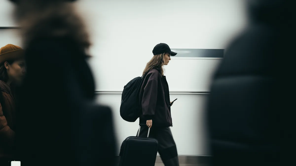 A young woman in a black cap and jacket walks with a backpack and suitcase, looking at her phone.