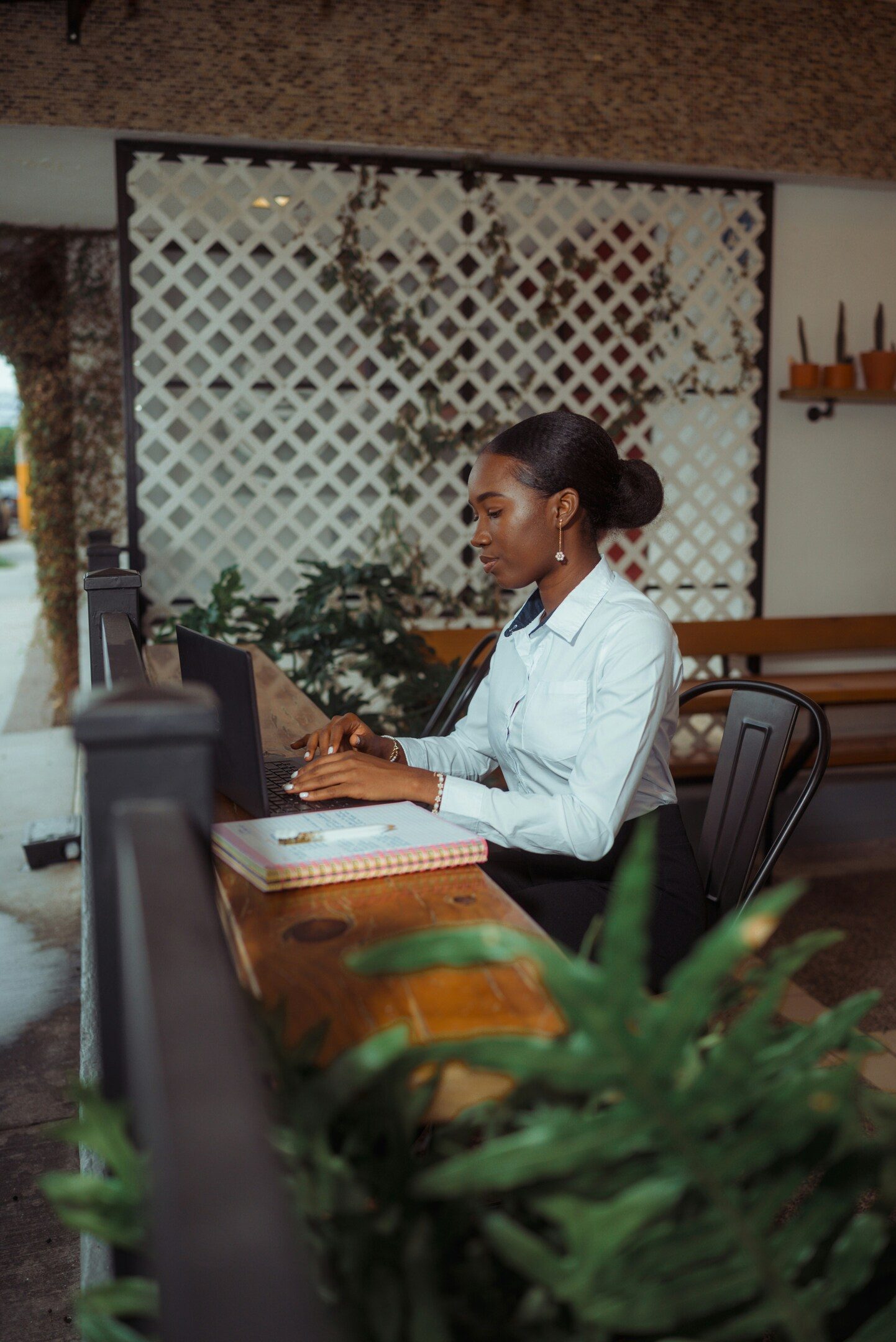 A woman in a white shirt works on a laptop at a wooden table in an outdoor or cafe setting.