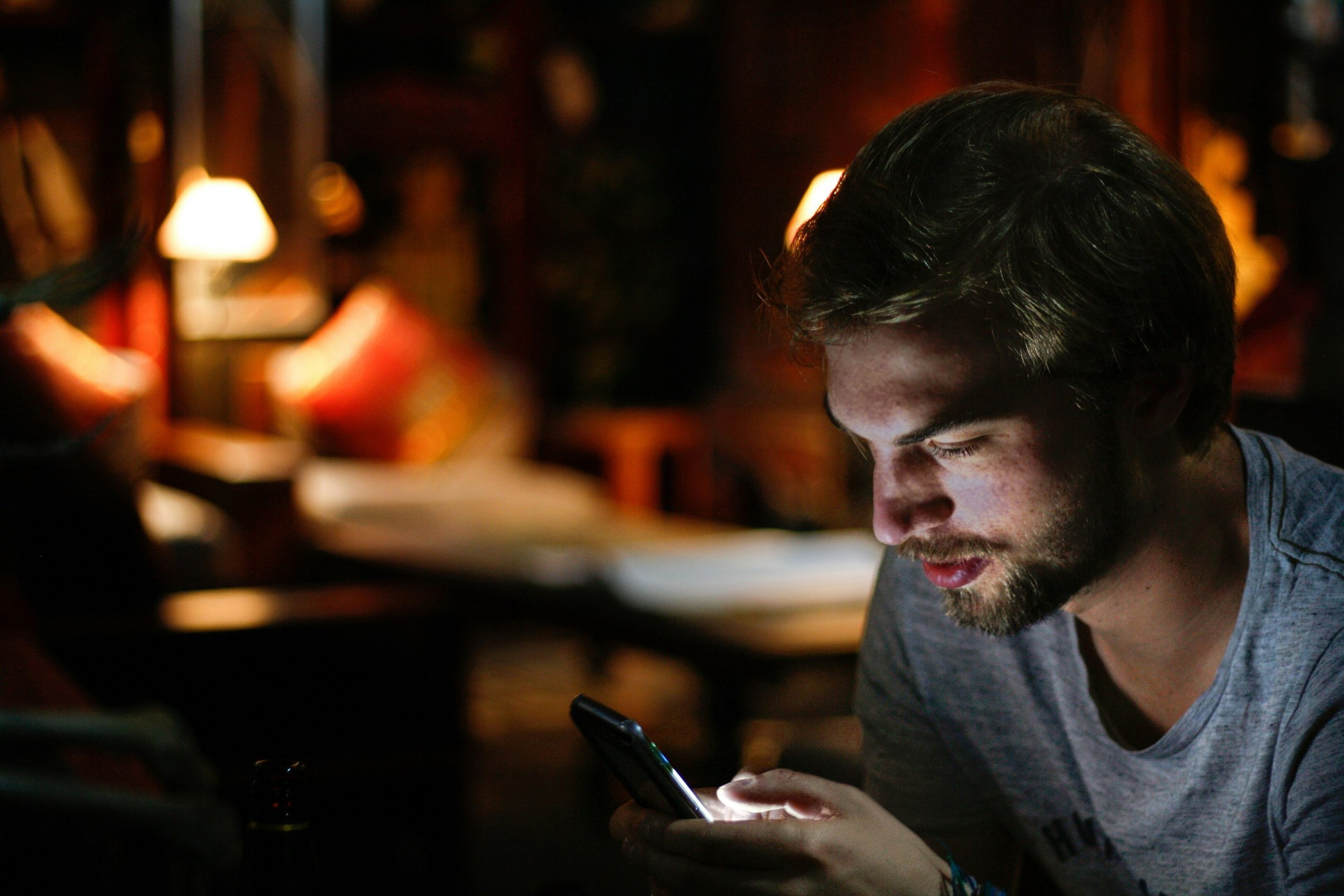 Young man with a beard looking intently at his glowing smartphone in a dimly lit setting.