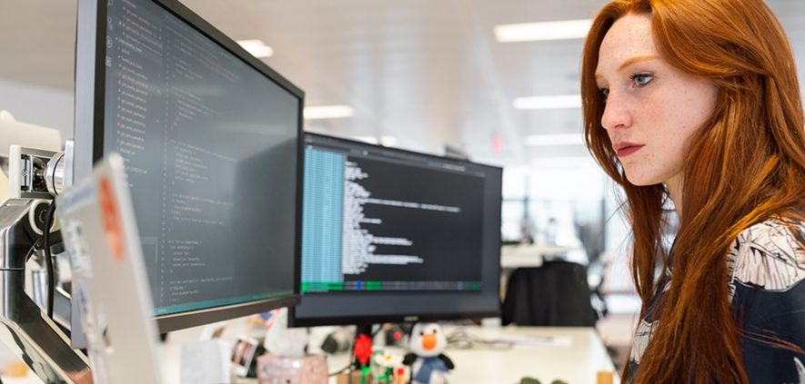 A focused woman with red hair coding at a computer workstation with dual monitors displaying code.